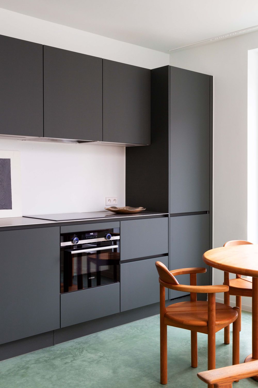 Modern minimalist kitchen with dark gray cabinets, built-in appliances, and a wooden chair on mint green flooring.