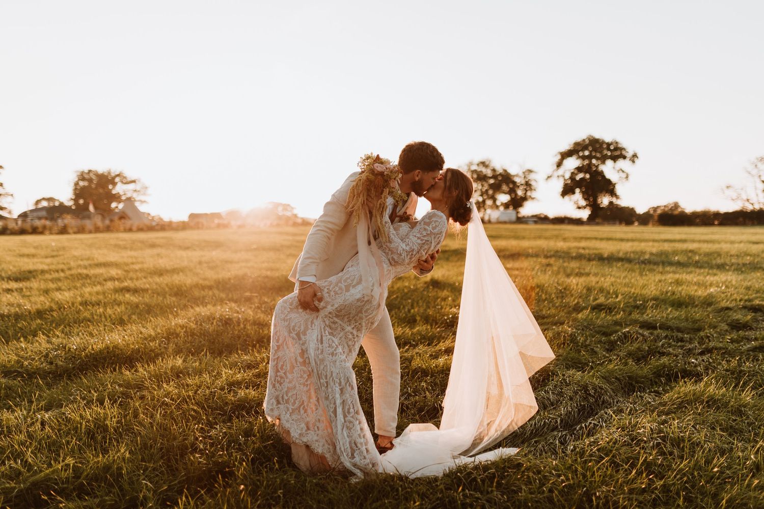 A couple shares a romantic moment in a golden sunlit field during sunset, with her white dress flowing in the breeze at Stanford Farm