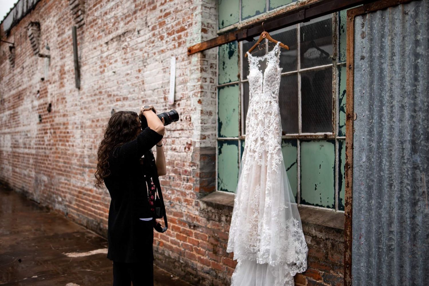 A photographer captures a detailed shot of an elegant lace wedding dress hanging against a rustic brick wall.