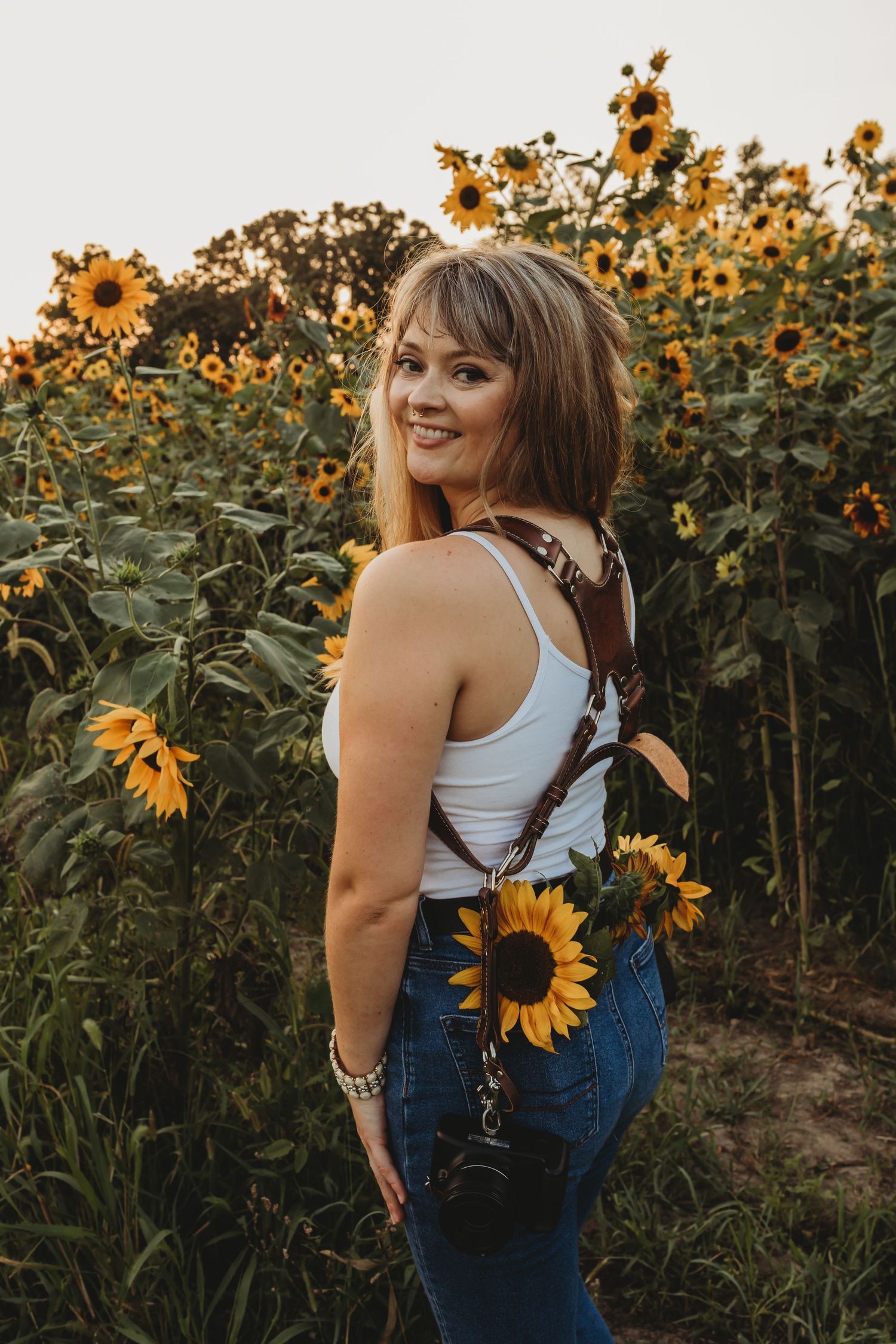 A photographer poses with sunflowers at sunset while wearing a white tank top and jeans.