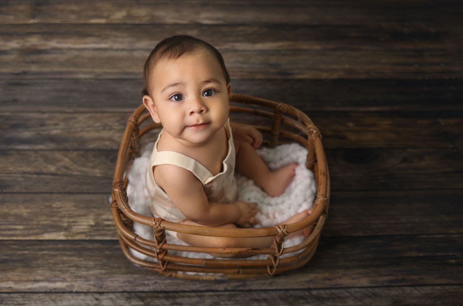 Baby sitting in wooden basket smiling at camera against dark wood background.