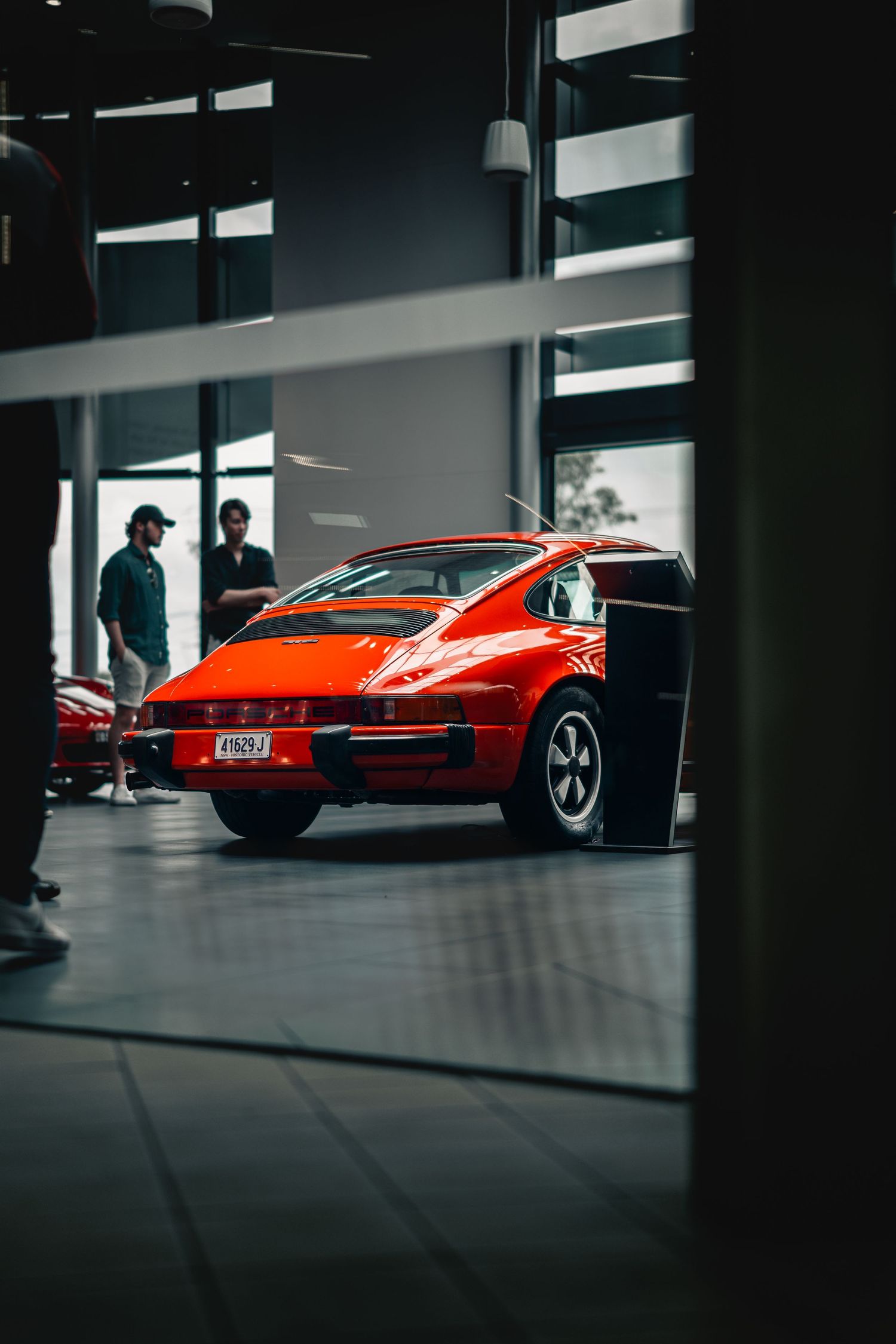 A red classic Porsche 911 seen through window blinds in a modern showroom setting.