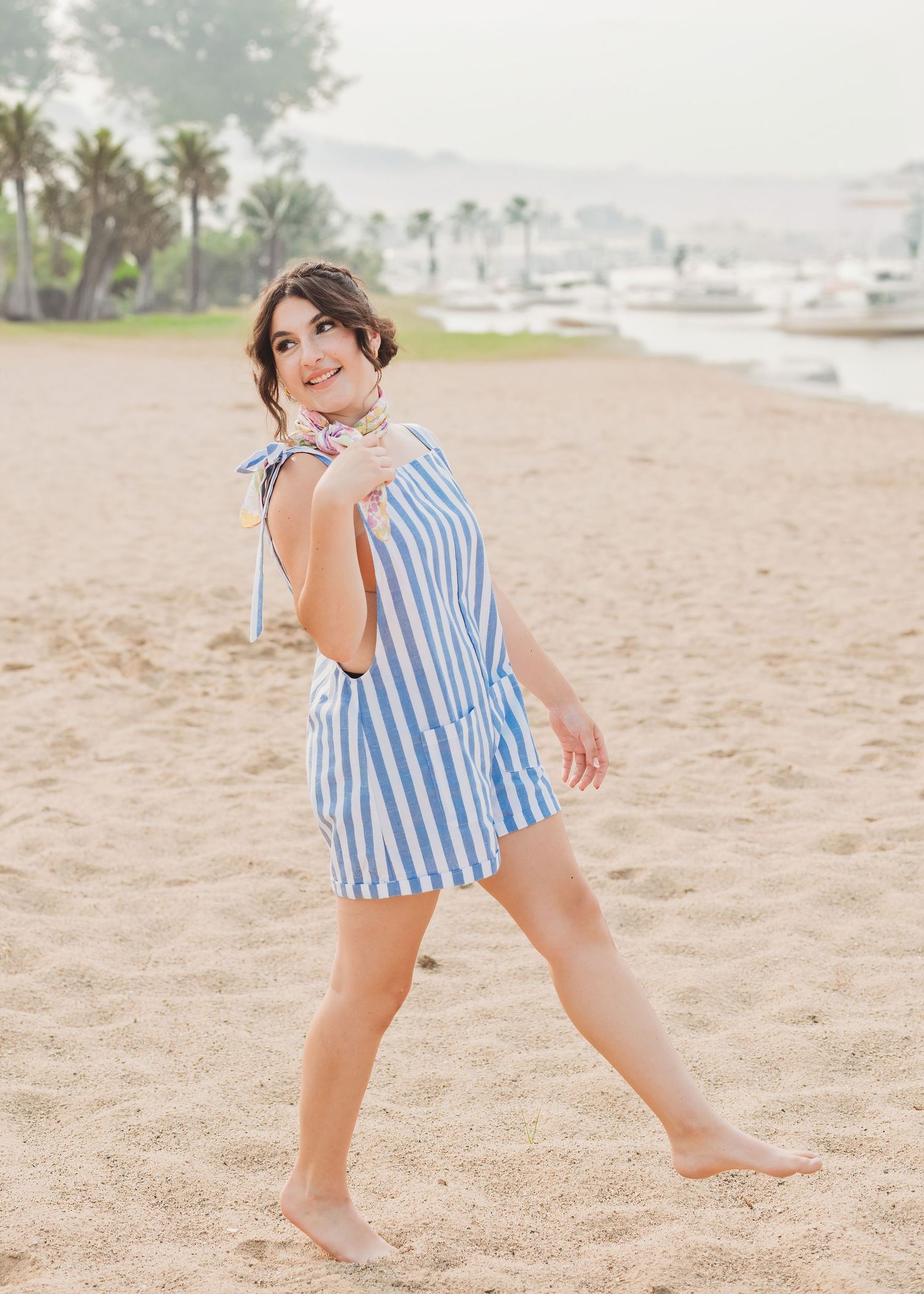 Woman in striped blue and white dress walking barefoot on beach with palm trees in background.