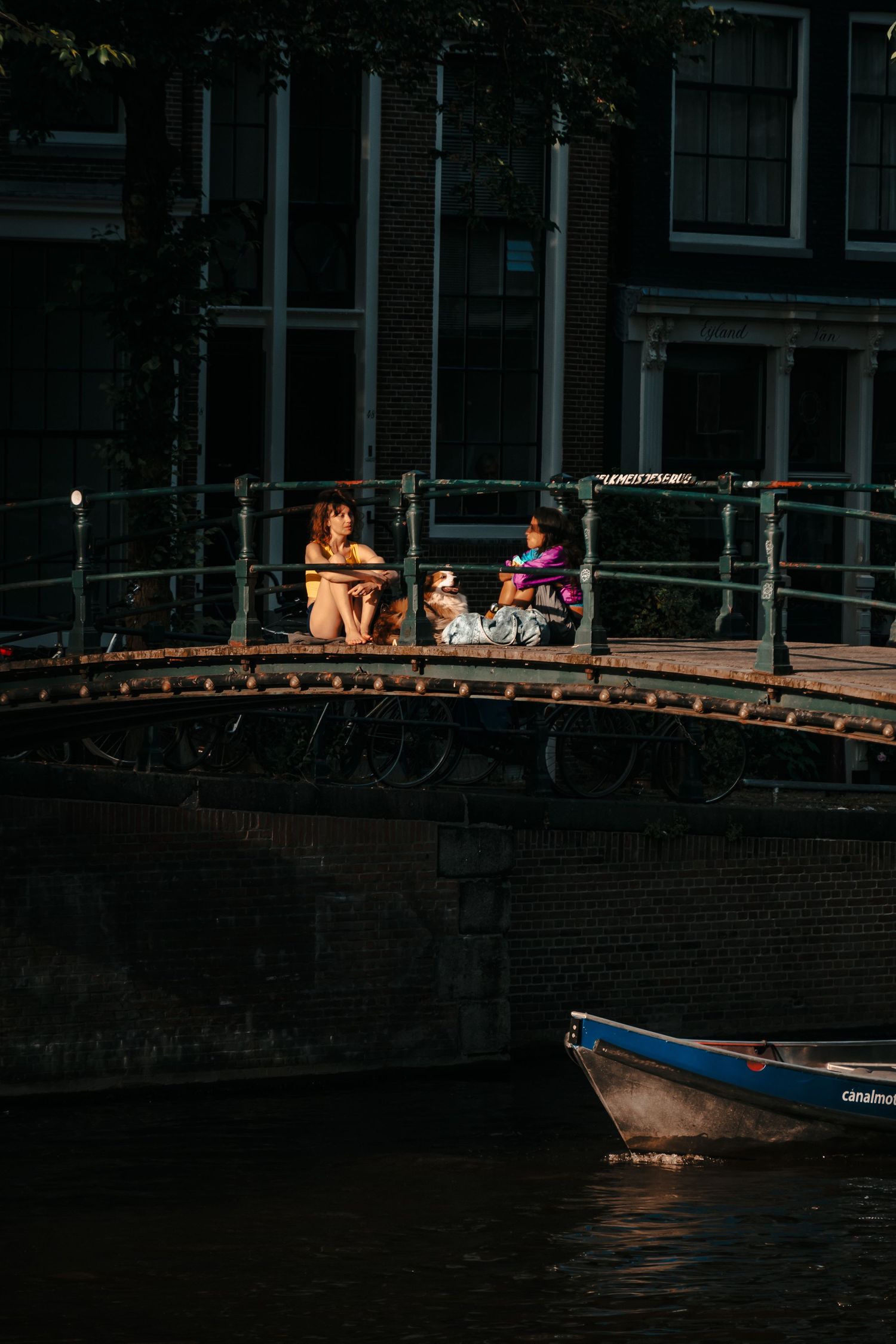 Two woman and a dog sitting on the Melkmeisjesbrug in Amsterdam center while a boat sails under them on a summer night.