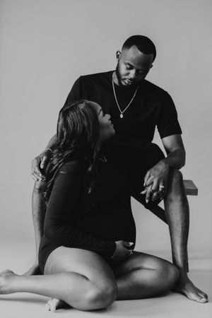 Black and white maternity photo of a couple, woman sitting on floor looking up at her partner.