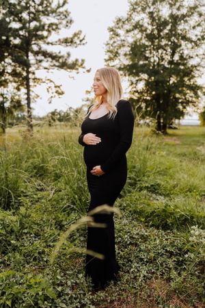 Pregnant woman in black dress cradling baby bump outdoors in lush green field.
