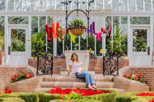 A one of a kind senior portrait. Girl sits on bricks in a conservatory filled with flowers. Vibrant colors, crisp, clean