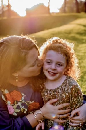 a mother gives her daughter a kiss on the cheek while sunlight shines behind them