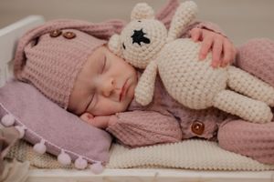 a sleeping baby holds a teddy bear posed on a tiny bed dressed in a night cap