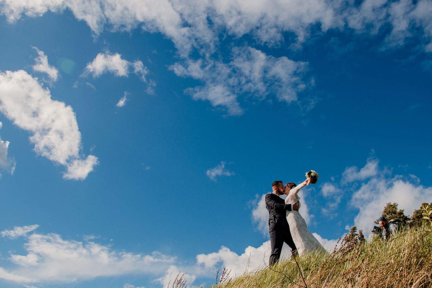 A joyful wedding couple silhouetted against blue sky and clouds on a hilltop.