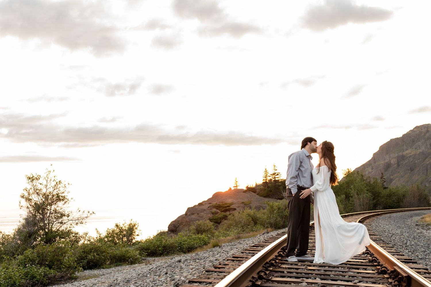 A romantic couple poses on train tracks at sunset with mountains in the background.