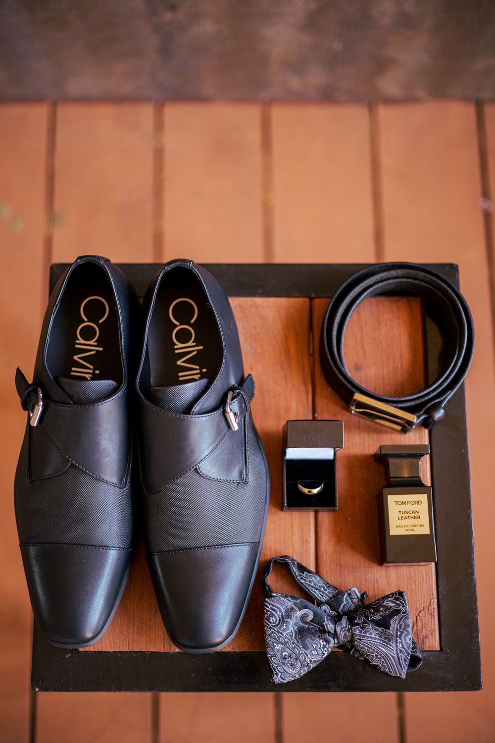 Groom's accessories including black dress shoes, bow tie, belt, cologne and ring box on wooden surface.