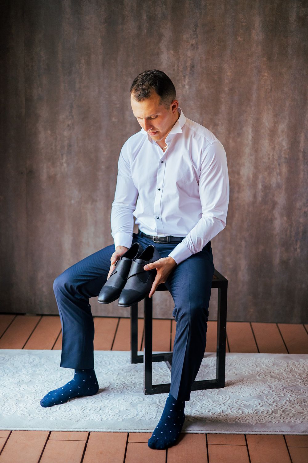 A person in business attire sits examining dress shoes on a black chair against a textured wall.