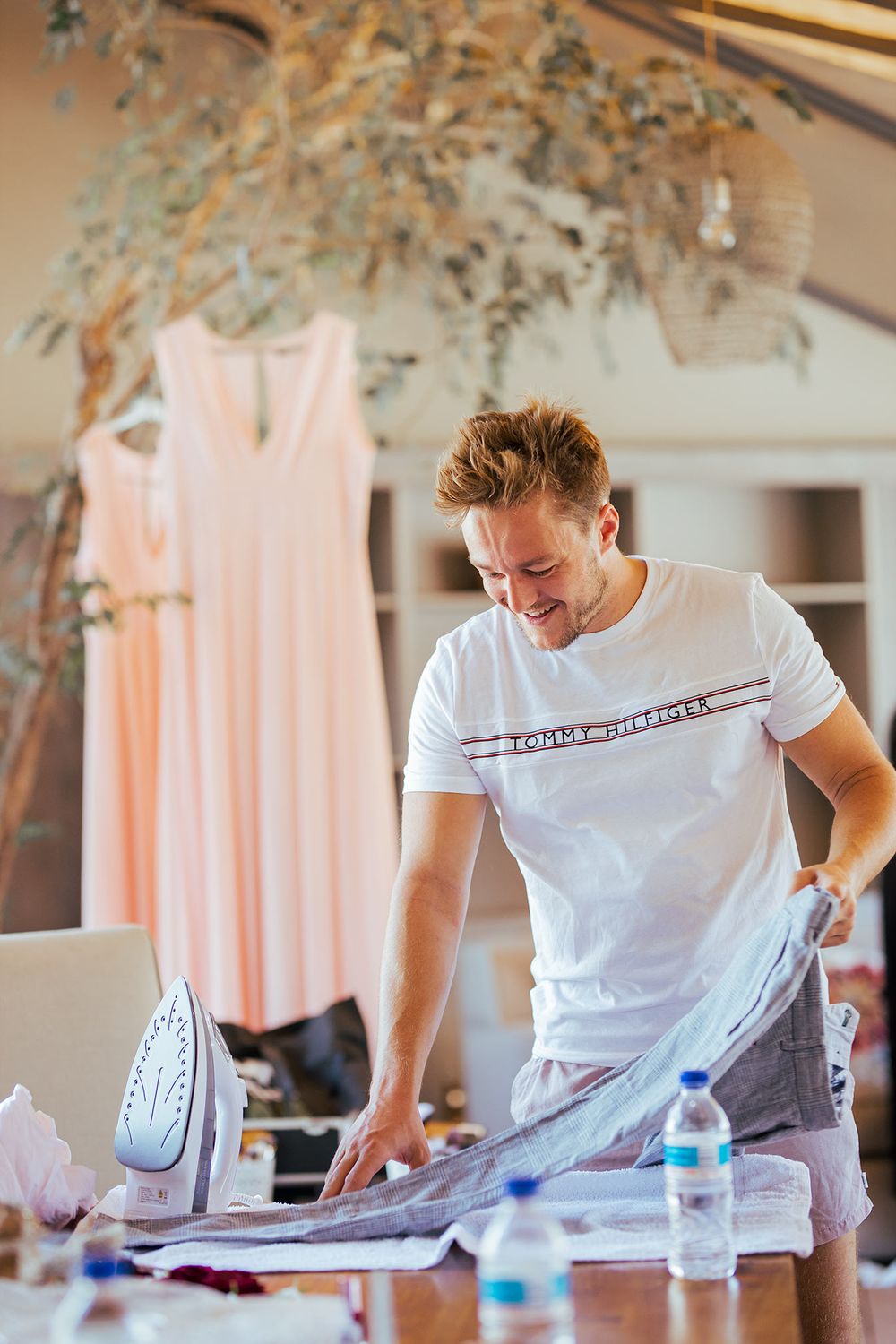 Person ironing clothes during wedding preparations in a room with dresses hanging in the background.