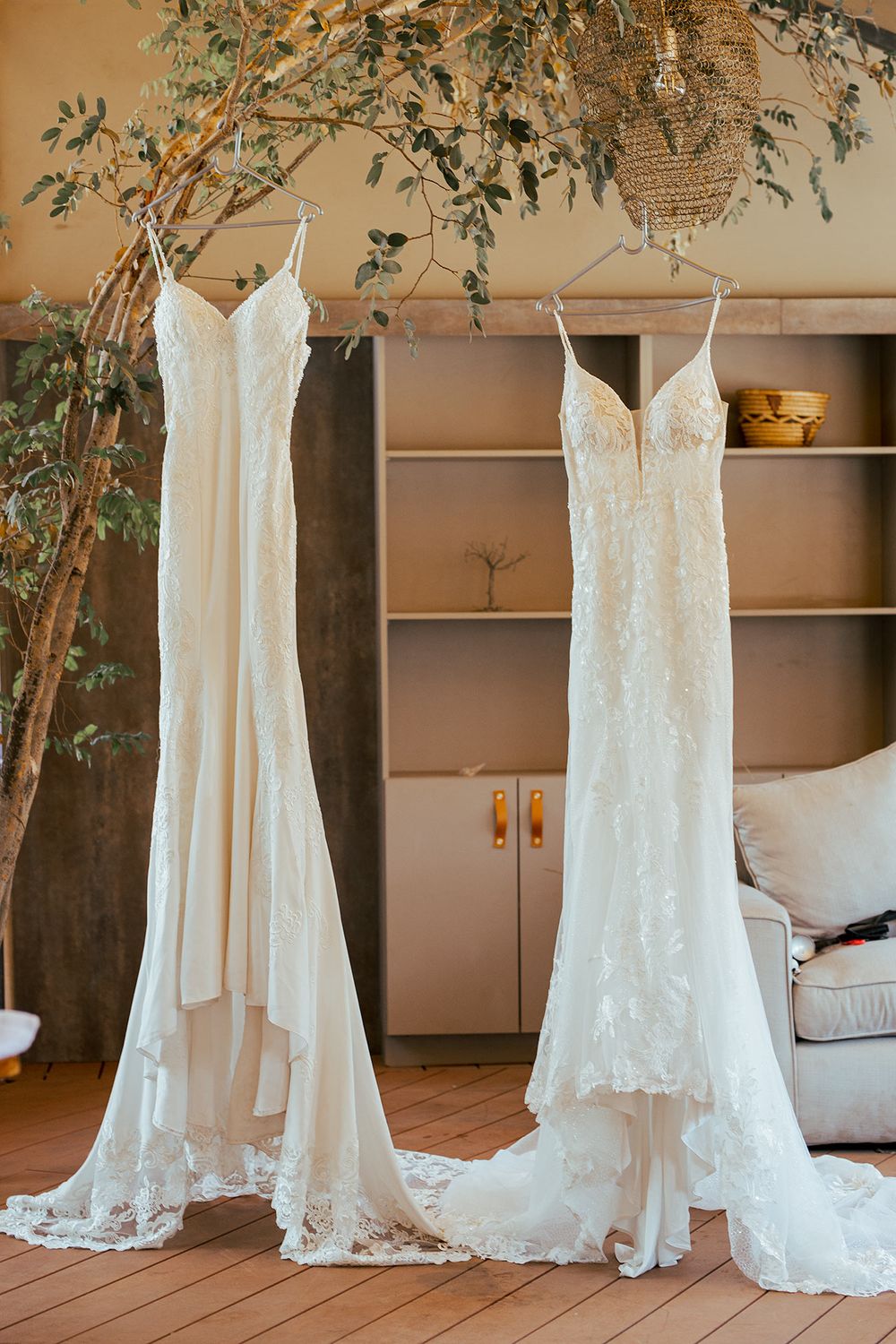 Two elegant white lace wedding dresses hanging on display with greenery decor overhead.
