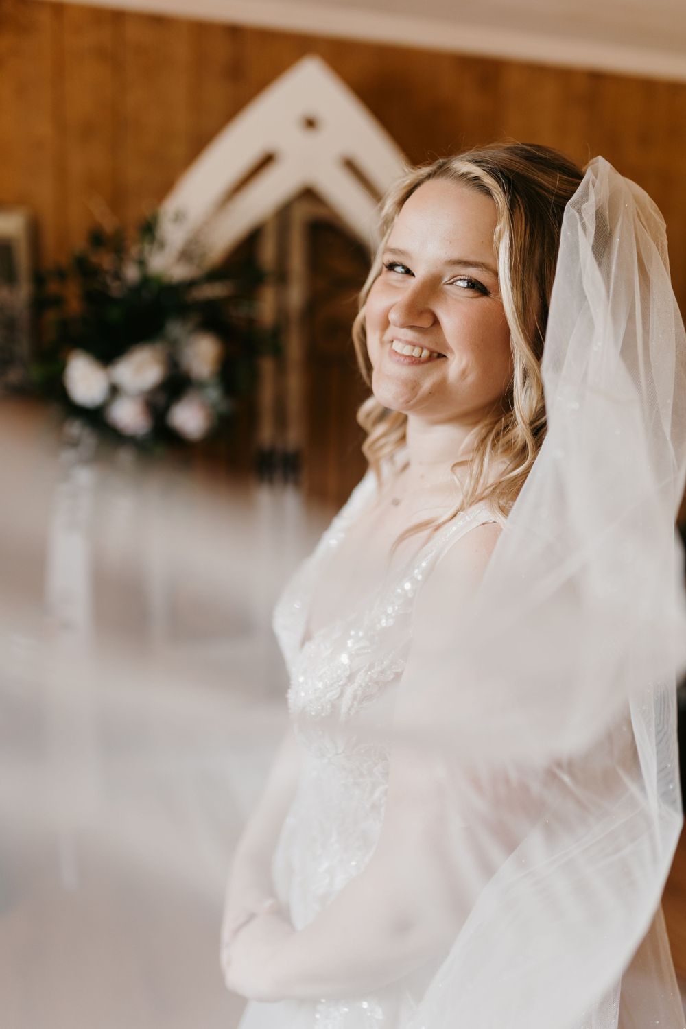 Radiant smiling bride in lace gown and veil poses in rustic chapel-style wedding venue interior.