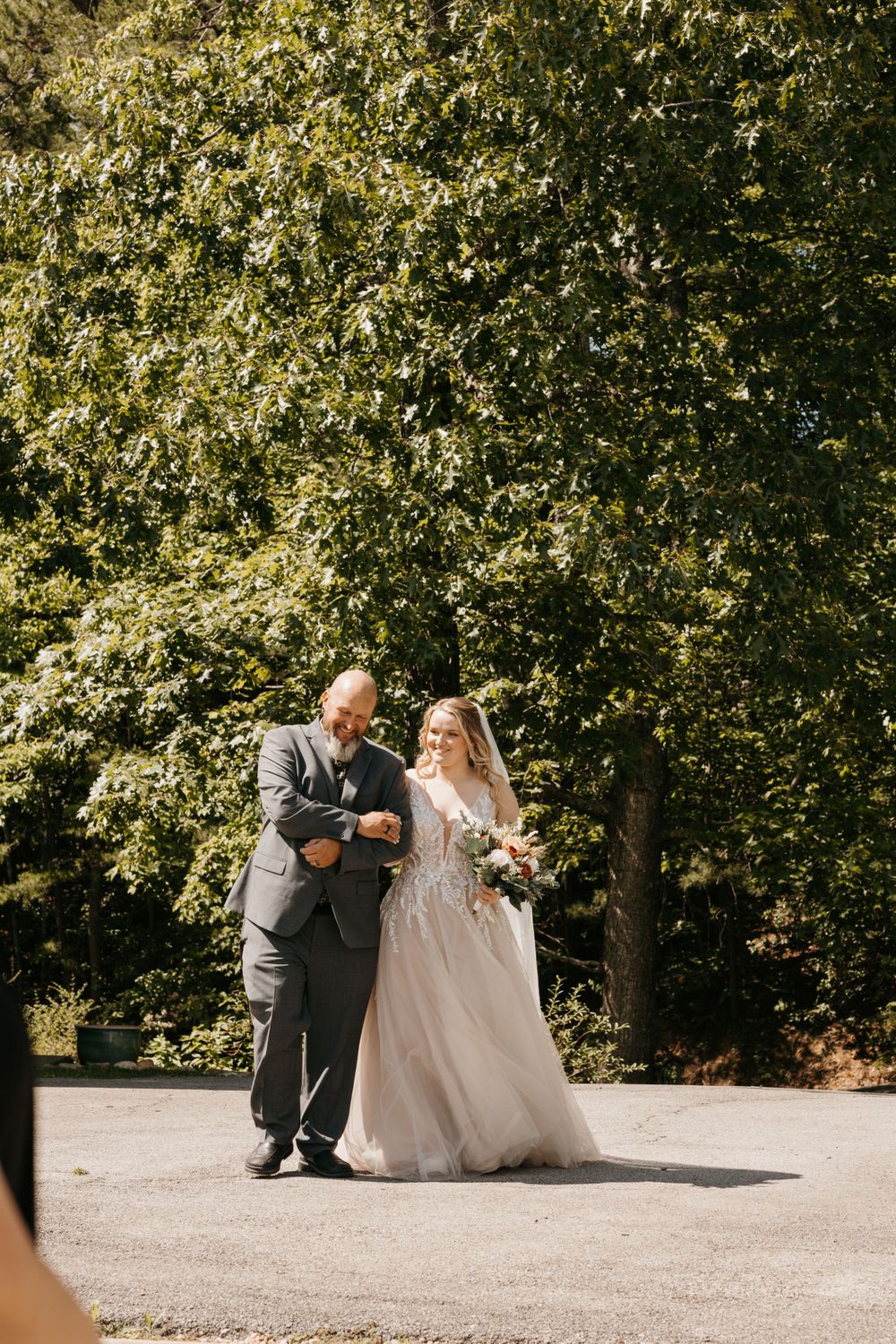 Bride in elegant lace gown walks with her father toward the wedding ceremony aisle surrounded by lush green trees.