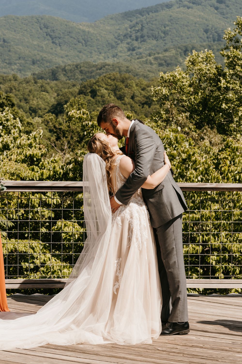 Newlyweds share first kiss on wooden deck with lush green mountain landscape in the background.