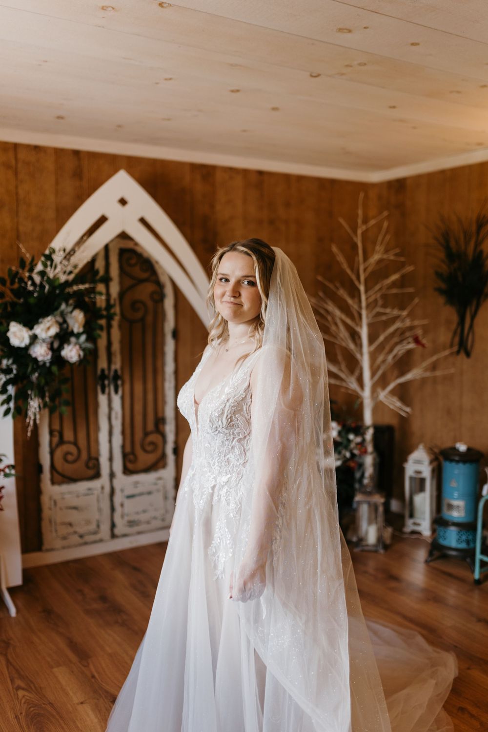 Elegant bride in lace wedding gown and cathedral veil poses in rustic wood-paneled wedding venue.