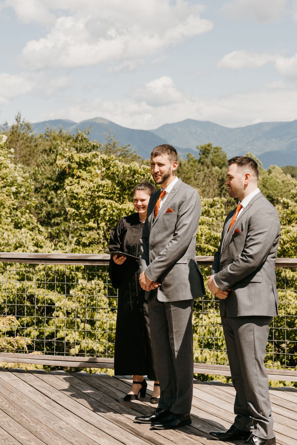 Groom and groomsman in gray suits with orange ties stand at mountain overlook wedding ceremony with officiant.