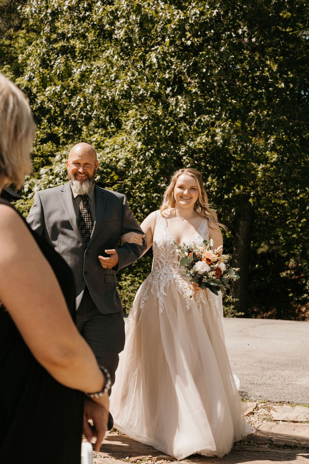 Smiling bride carrying floral bouquet approaches guest while escorted by bearded man in gray suit.