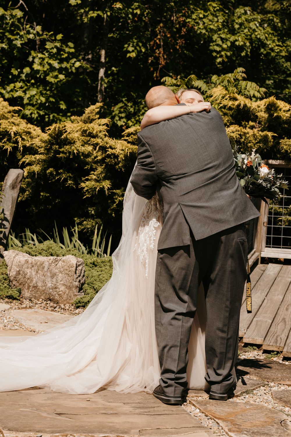 Bride in lace wedding gown shares emotional hug with escort on stone patio surrounded by greenery.