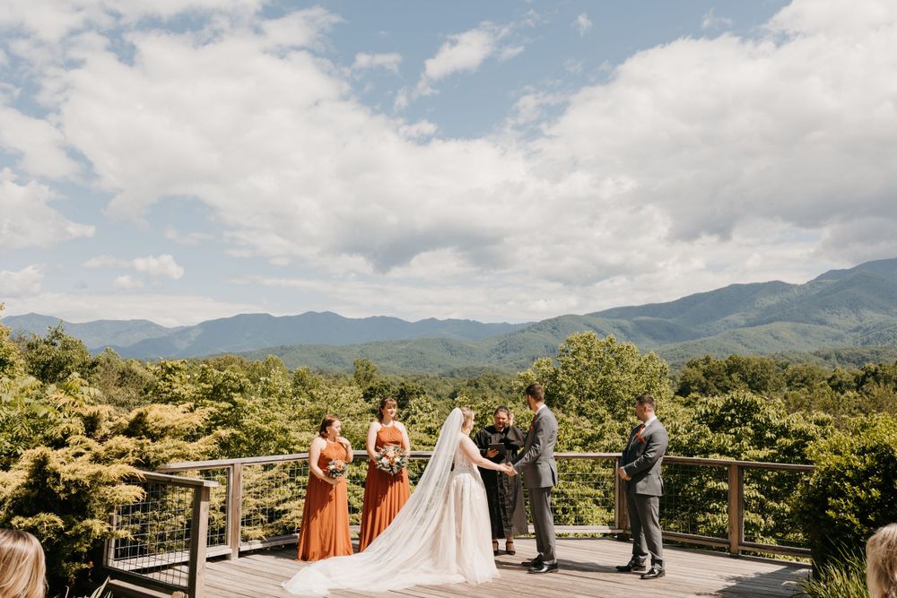 Bride and groom exchange vows on a wooden deck with breathtaking mountain landscape backdrop and wedding party.