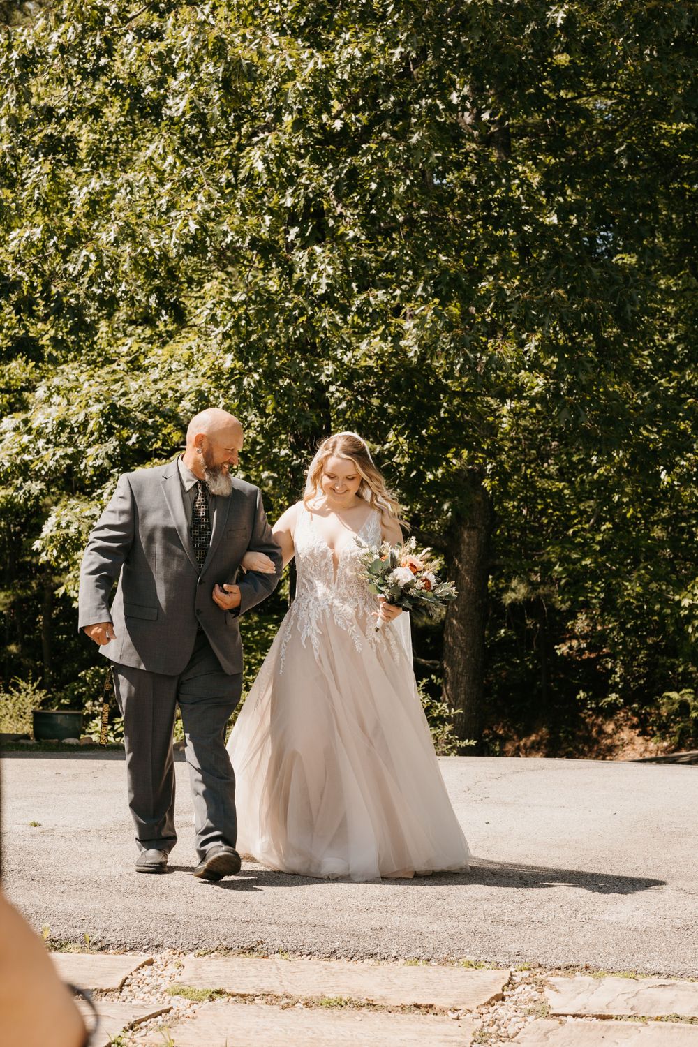 Bride in white tulle gown walks with bald bearded man in gray suit toward wedding ceremony outdoors.