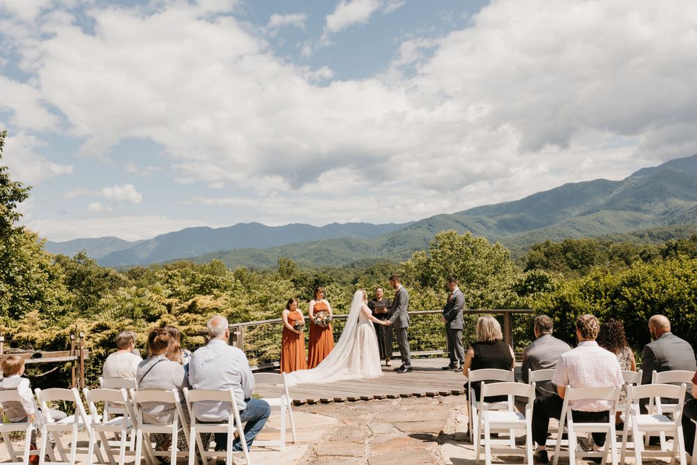 Outdoor mountain wedding ceremony with guests seated in white chairs on stone patio with panoramic mountain views.