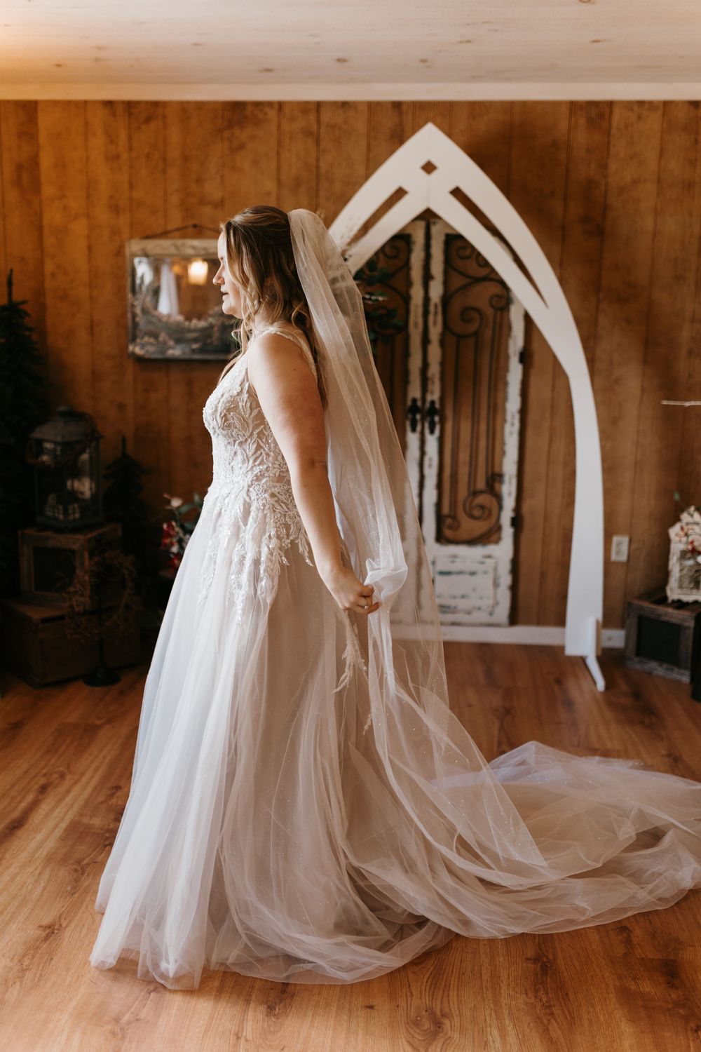 Bride in lace tulle wedding gown holds veil while posing in rustic wood-paneled venue interior.