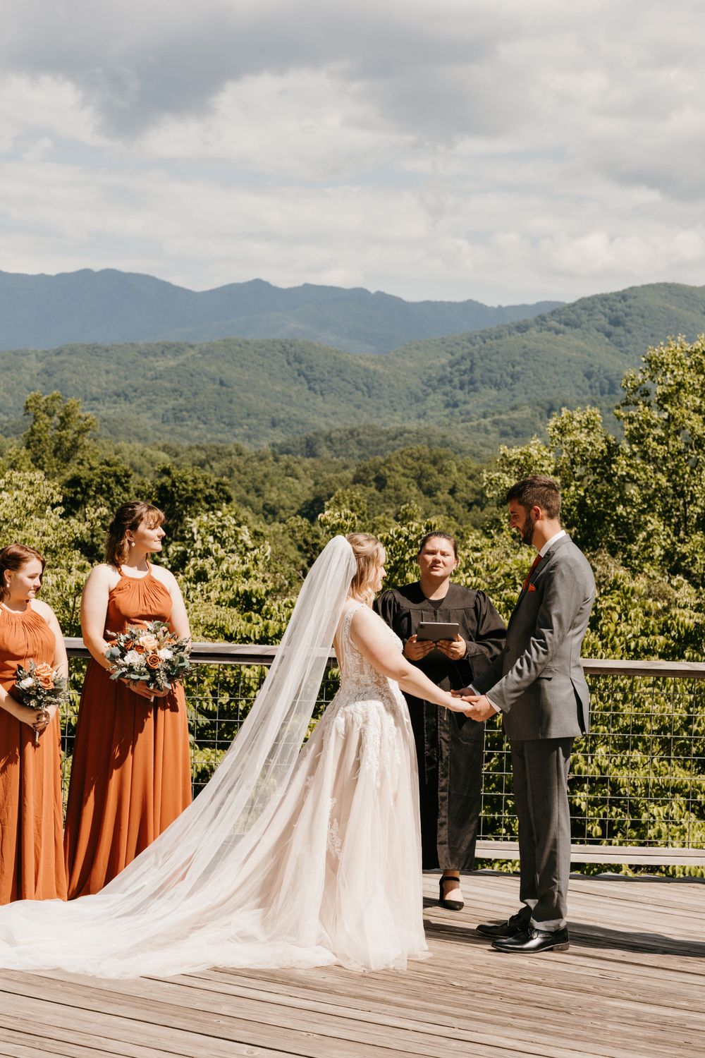 Bride and groom exchange vows on mountain overlook deck with bridesmaids in rust dresses nearby.