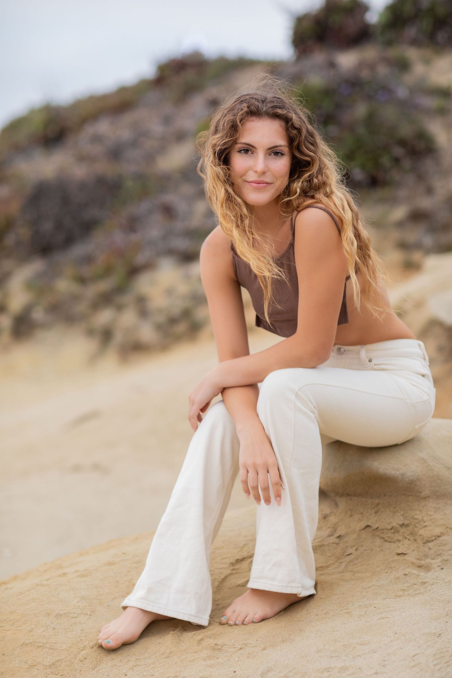 A high school senior in casual attire sitting on sandy beach cliff during golden hour photoshoot.