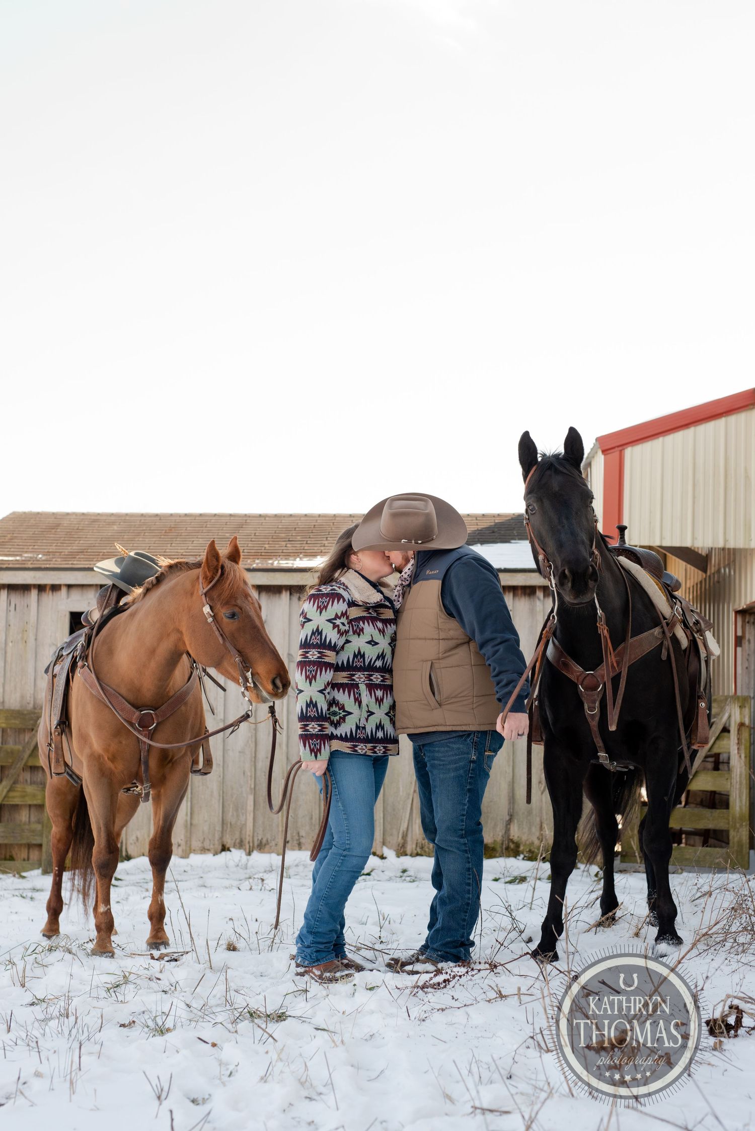 Two horses stand in the snow next to riders near farm buildings on a winter day.