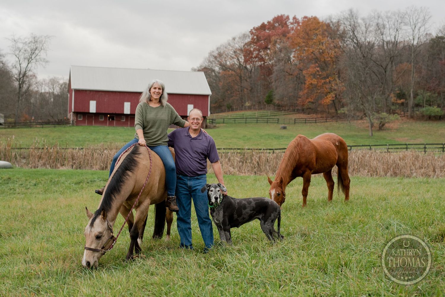 A couple with their black dog stands between grazing horses near a red barn in autumn.