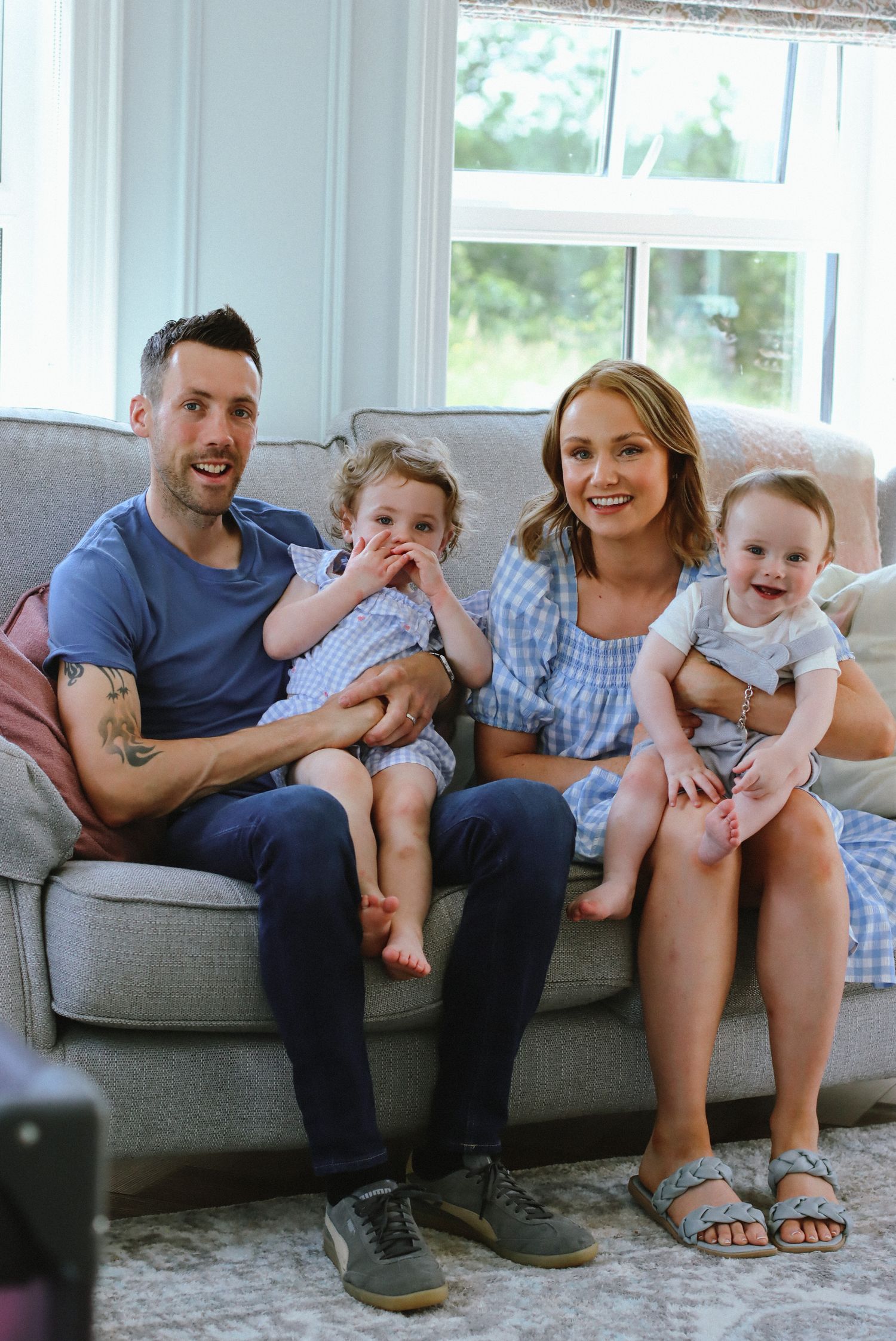 Family with two small children sitting together on a gray couch in a bright living room.