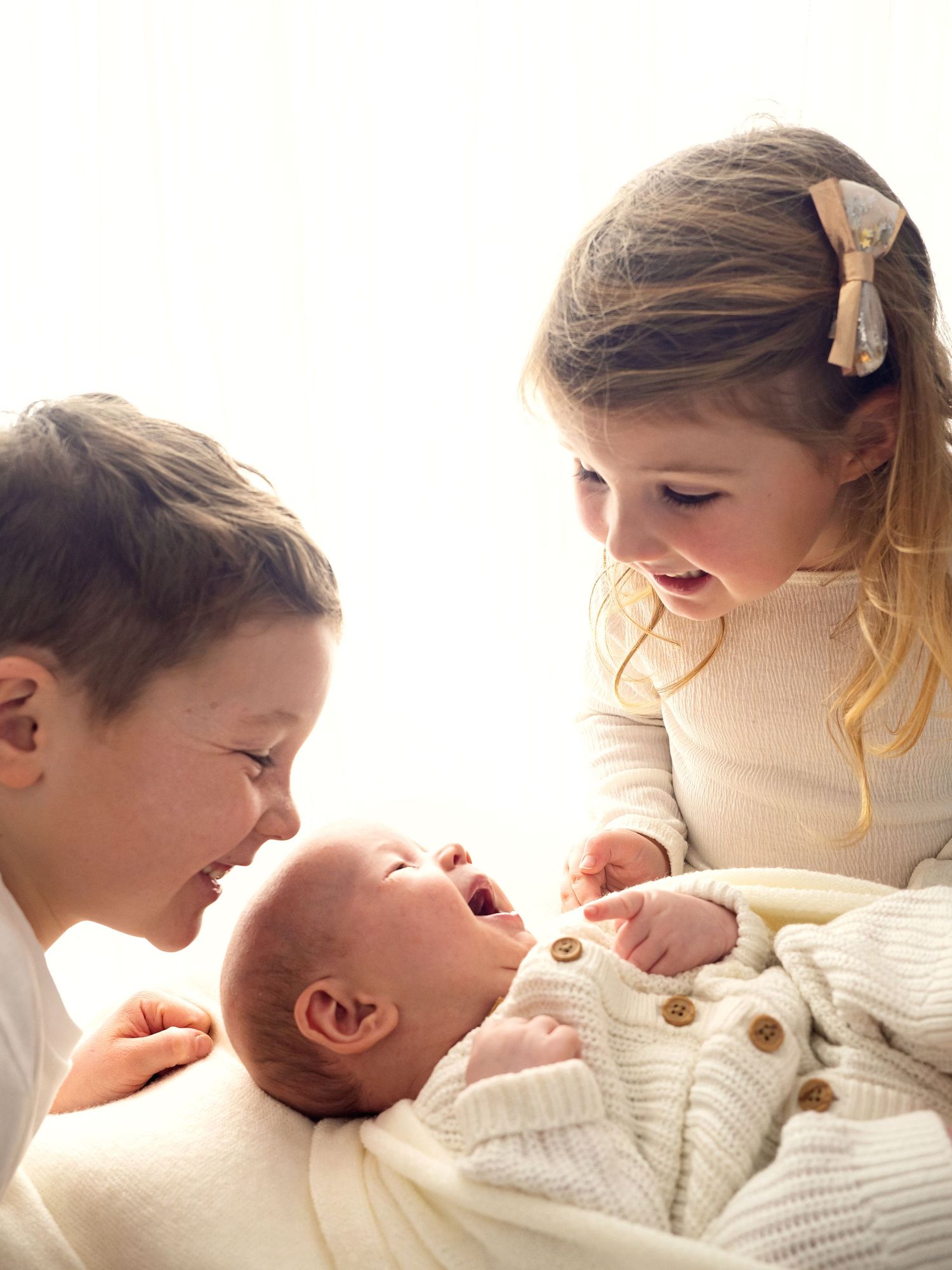 A young brother and sister smile and laugh looking at their newborn baby, during a photo session in Didcot