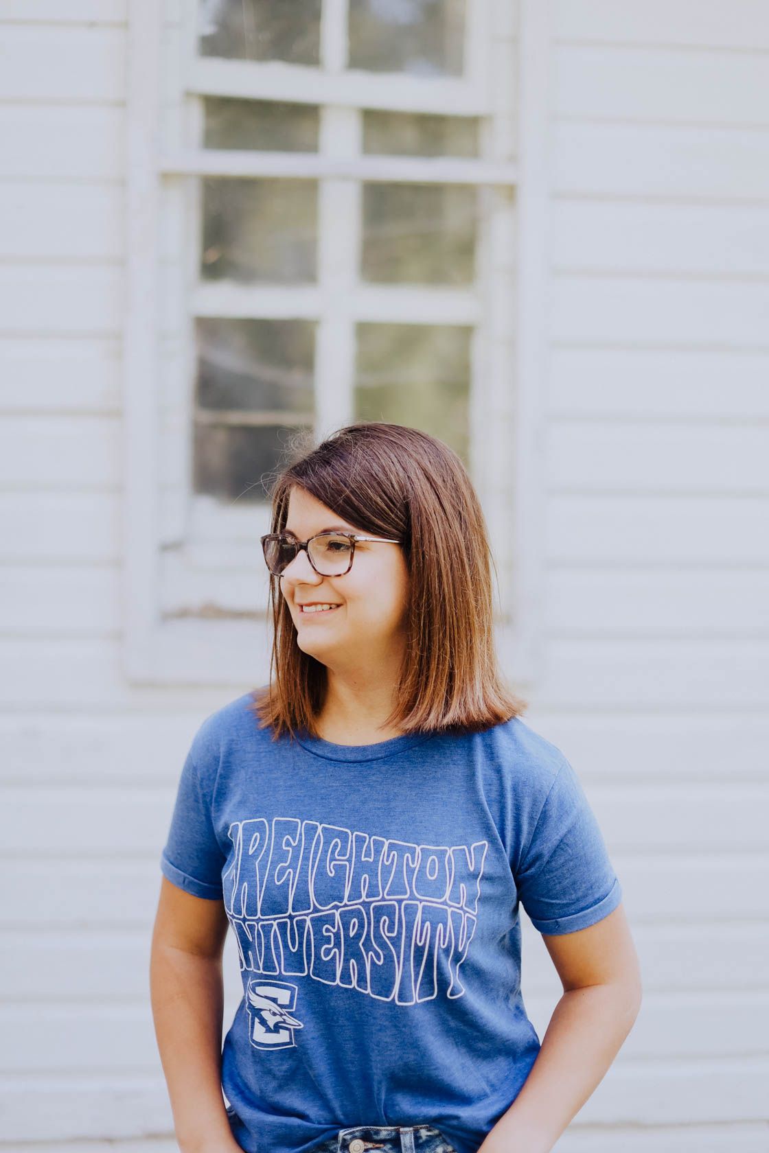 Person wearing glasses and a blue university t-shirt smiling against a white building exterior.
