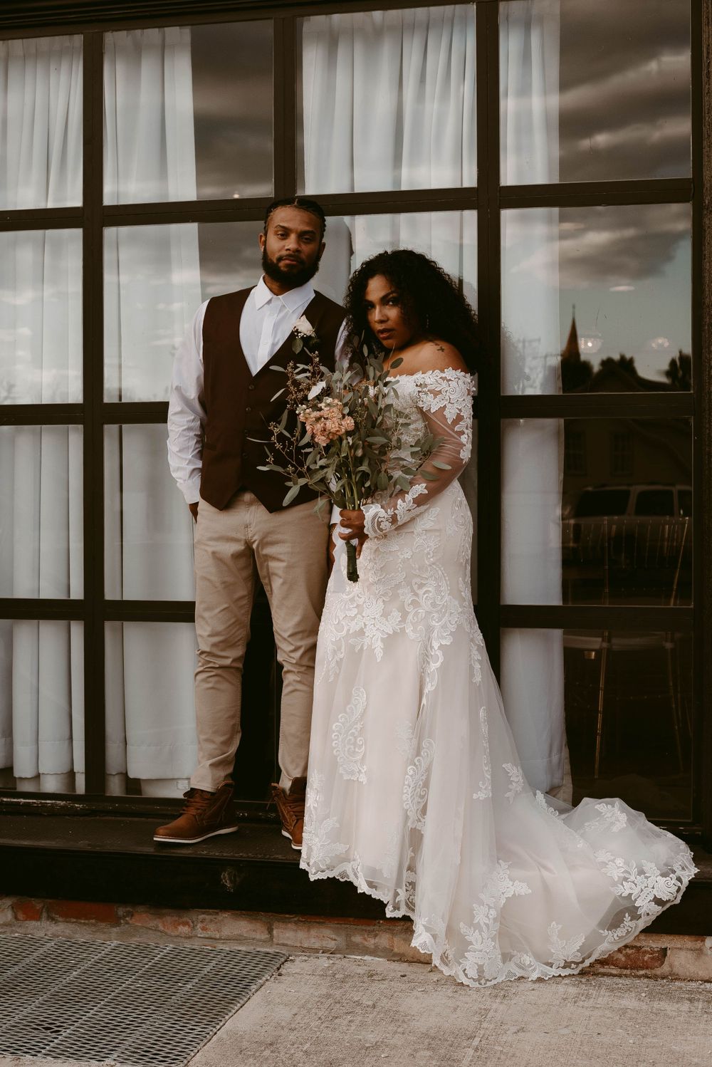 Couple in wedding attire stands against dramatic sky and window backdrop.