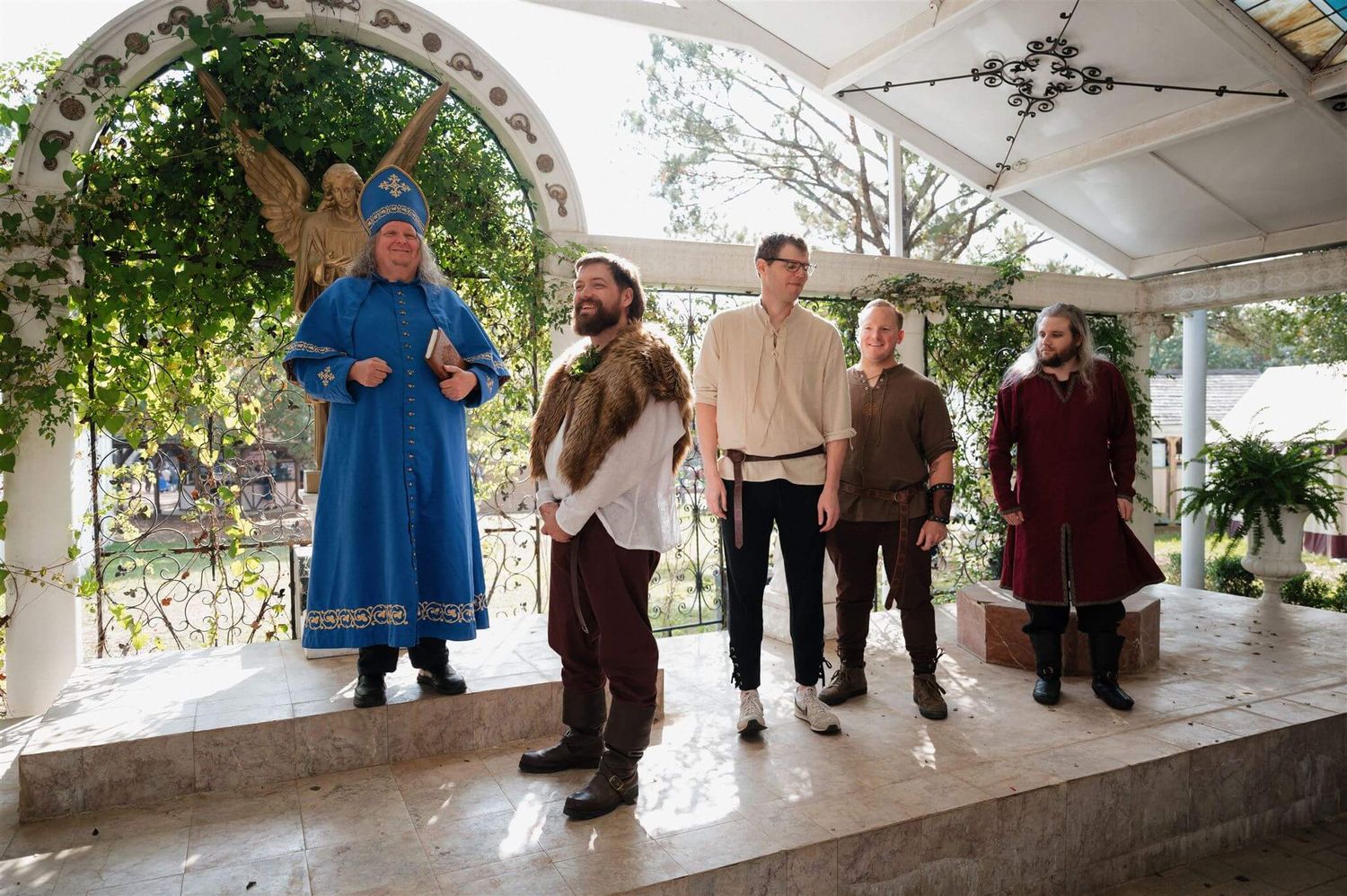 Group in period costumes posing on stage at renaissance fair under decorated white archway.