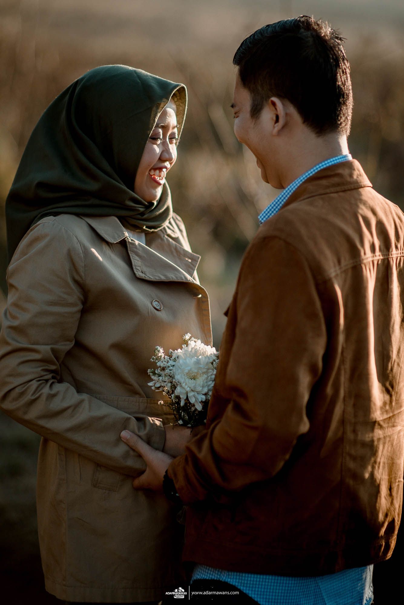 Muslim couple in casual attire sharing tender moment while holding small bouquet outdoors.