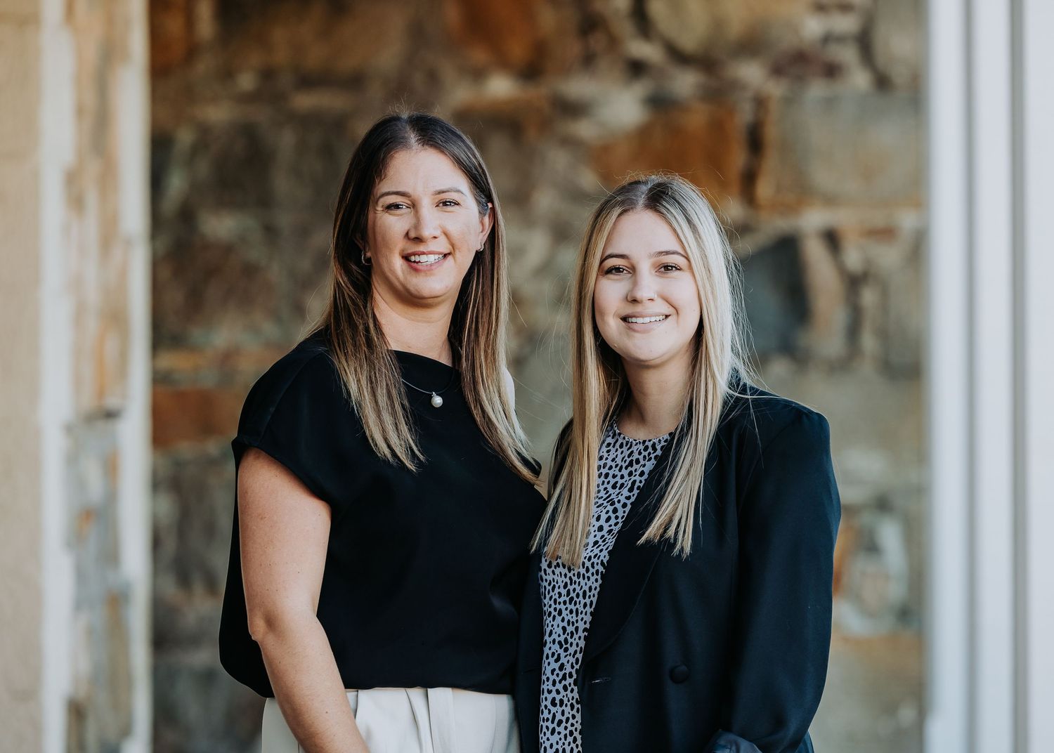 Professional photo of two colleagues in business attire standing together against a textured stone wall.