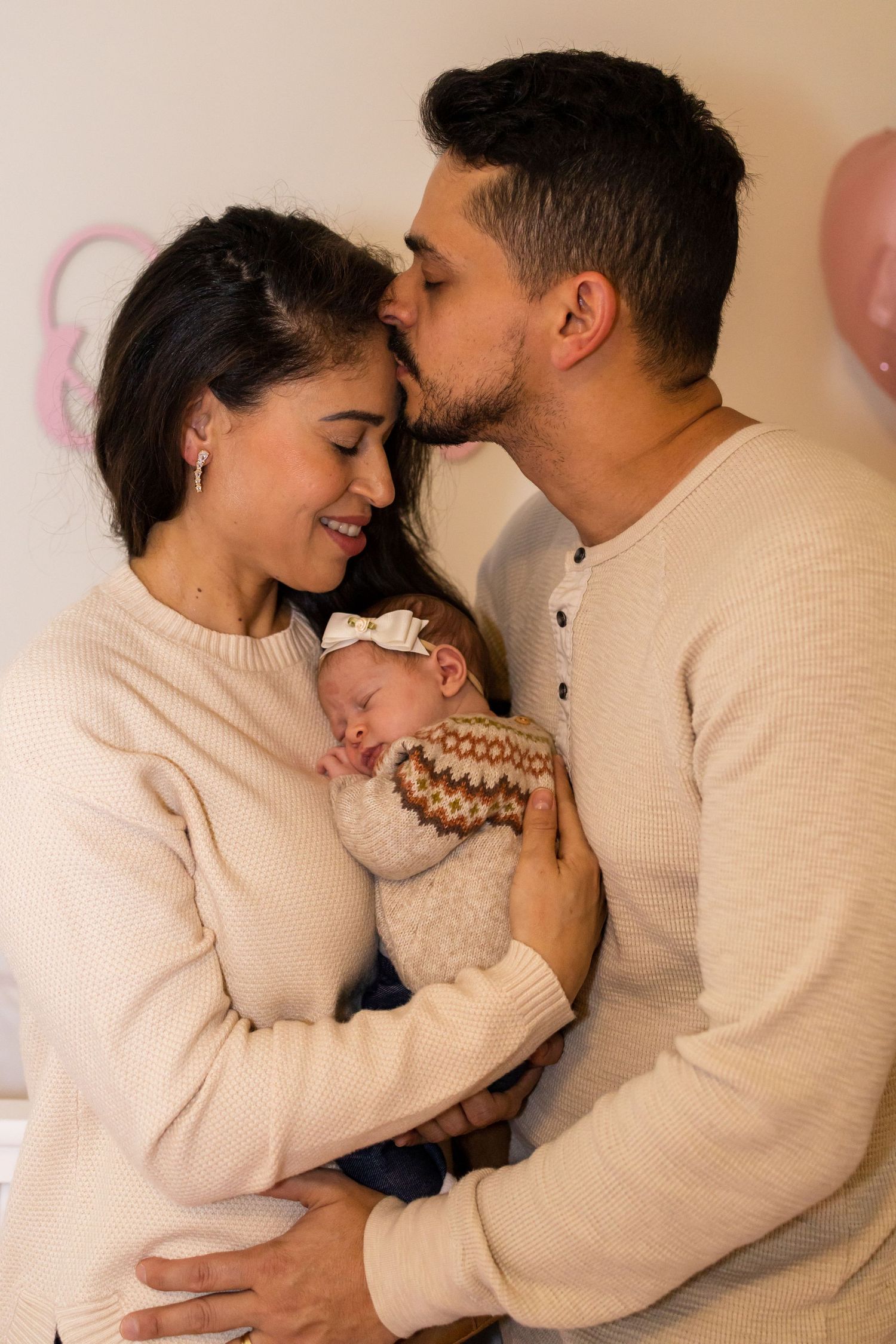 New parents in matching cream sweaters share tender moment with newborn baby.