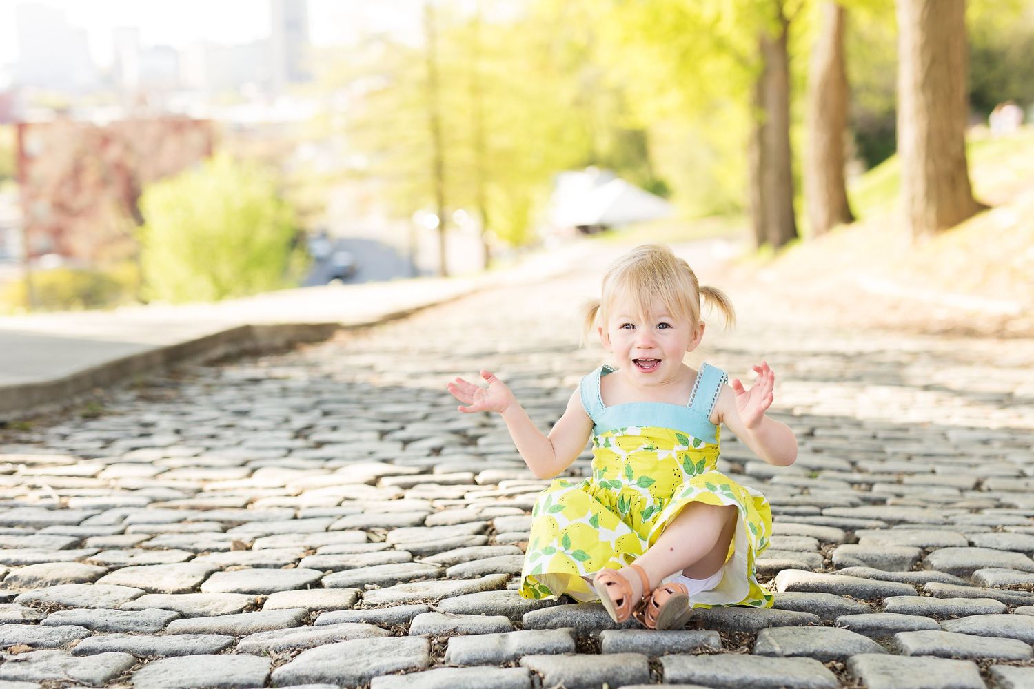 Happy toddler in yellow dress sitting on cobblestone street during golden hour.