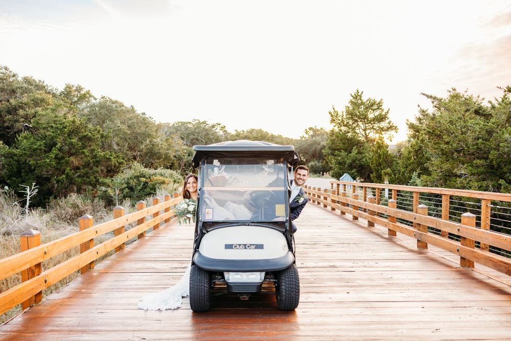 A golf cart drives across a wooden boardwalk bridge through a natural coastal landscape at sunset.