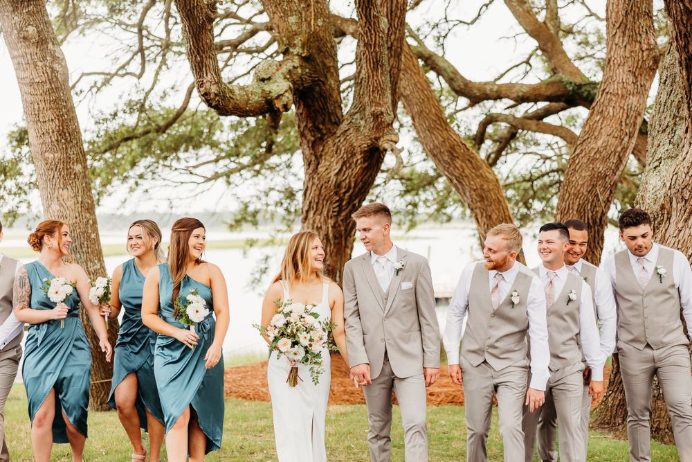 Wedding party in gray suits and blue dresses poses together under large trees at an outdoor venue during sunset.