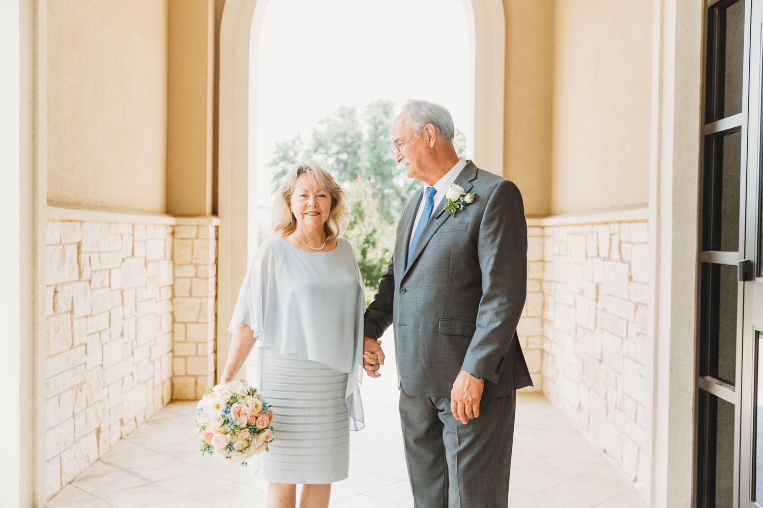 Elegant couple in light blue dress and grey suit pose along a stone columned hallway with beautiful floral bouquet.