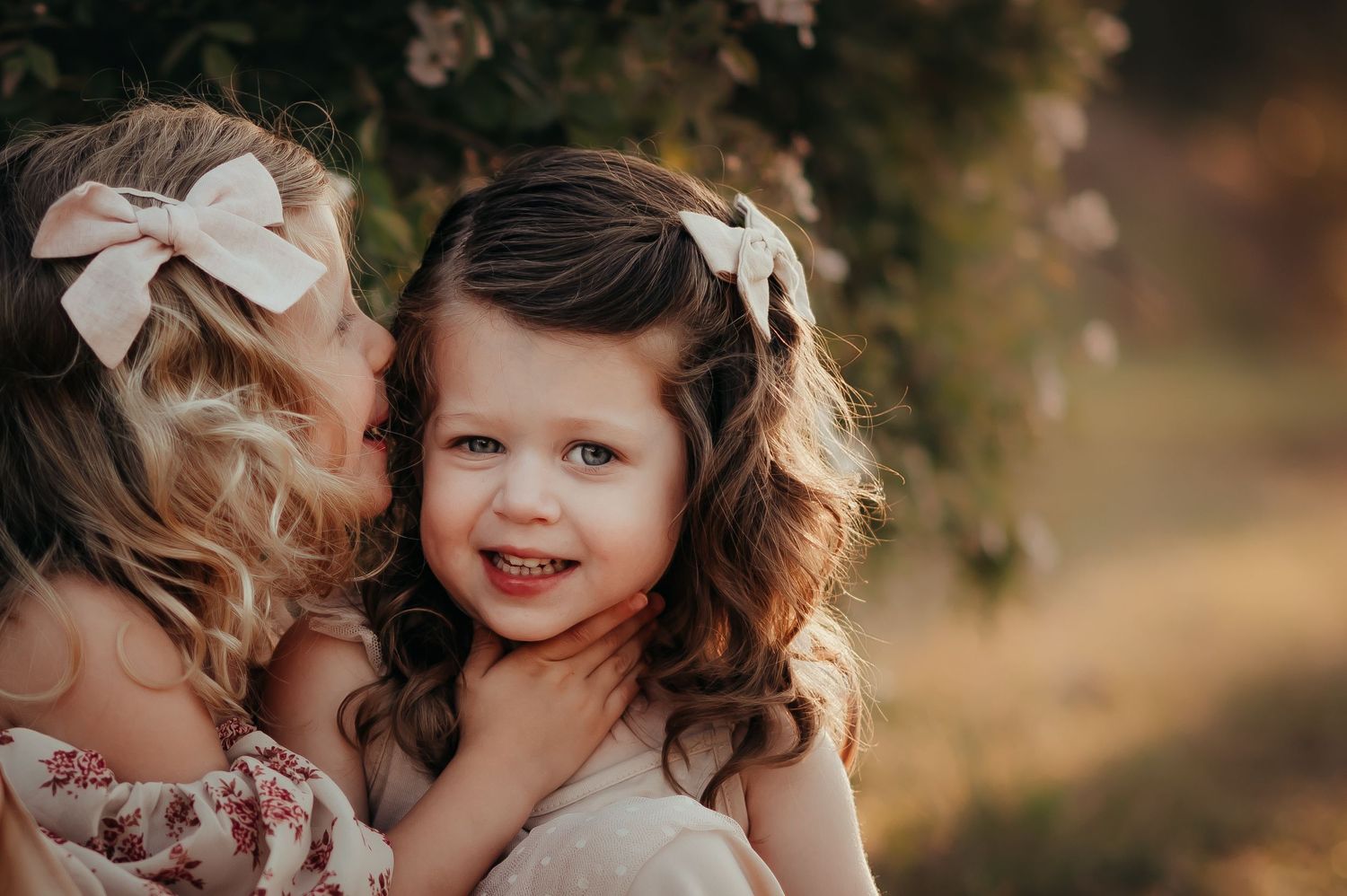 little blonde girl with bow and sitting by a bush tells a secret to her little cousin with brown hair and a bow.