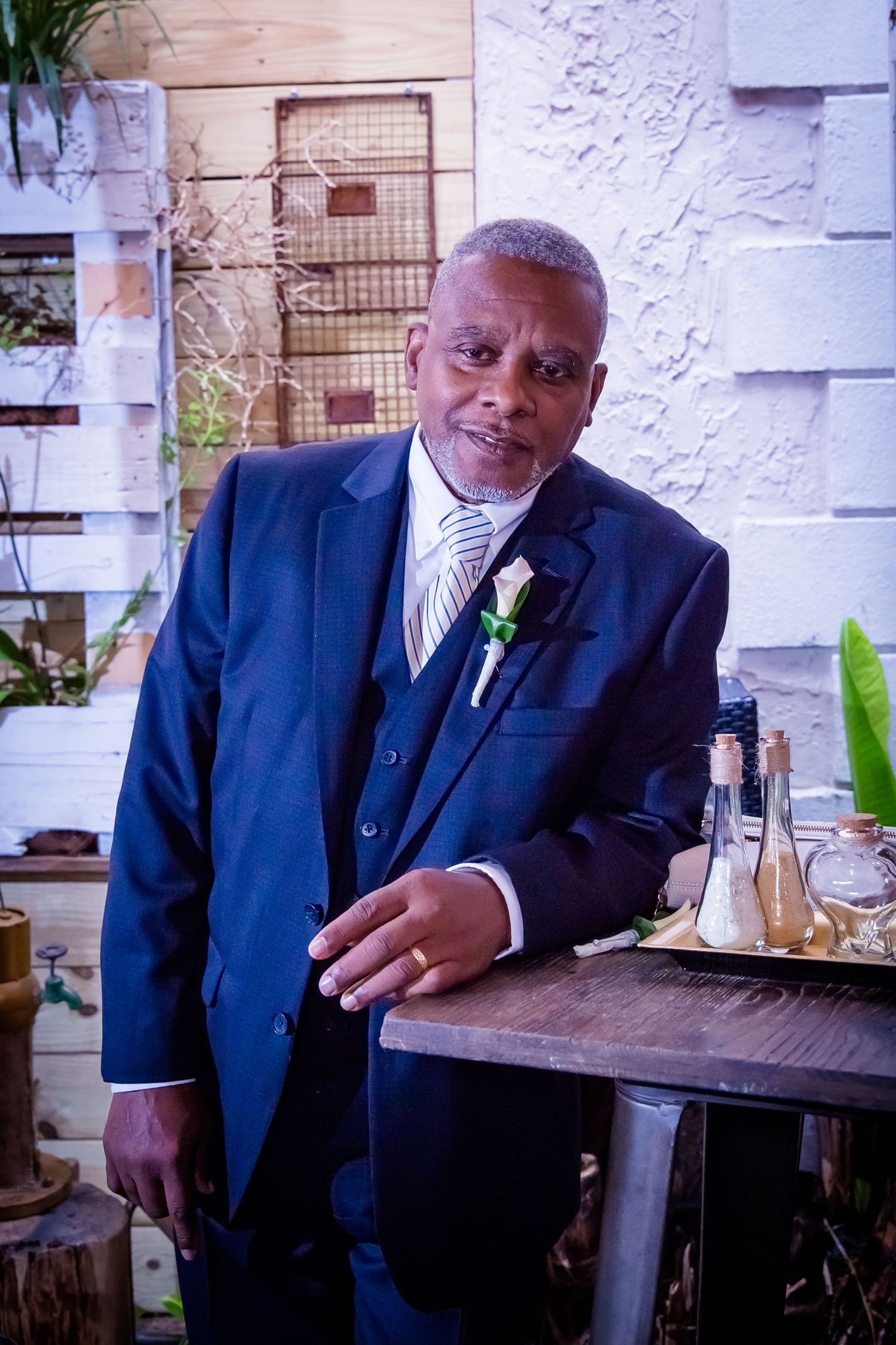 A person in a navy suit seated at a wooden table in an outdoor setting.