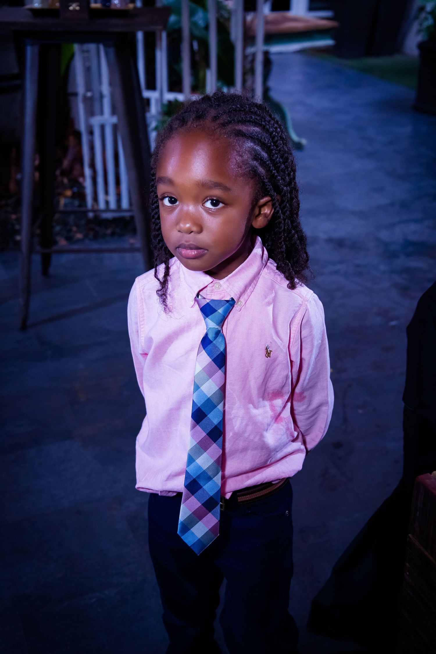 Young child wearing a pink shirt and plaid tie in a dark indoor setting.