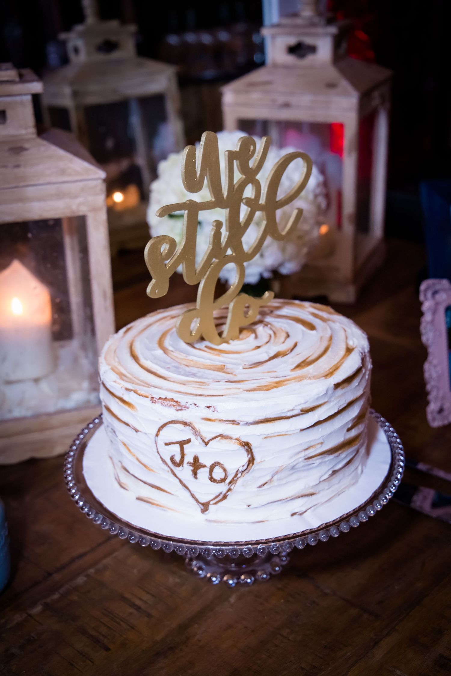 A rustic white wedding cake with gold 'We Still Do' topper surrounded by lanterns.